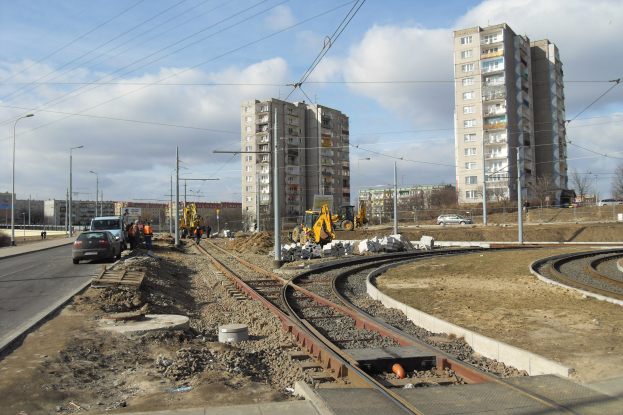 Baustelle mit einer Bahnschiene, Geb├Ąuden, Stra├čeninfrastruktur, Fahrzeugen, Passanten, Schutt, B├Ąumen und bew├Âlktem Himmel.