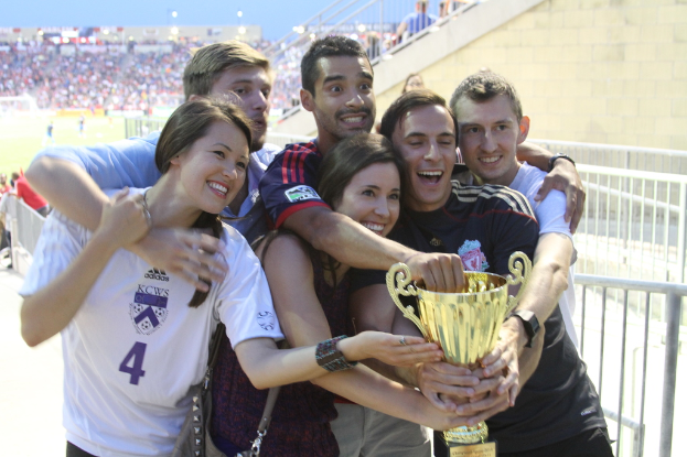 Sechs Menschen, zwei Frauen und vier Männer, halten einen Pokal und lächeln, posieren für ein Foto vor einem Eisengeländer, mit einer Wand und einem Geländer oben rechts, Menschen und Lichter im Hintergrund, möglicherweise in einem Stadion.