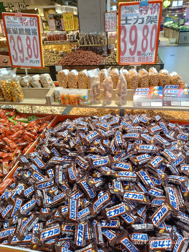 Eine Vitrine in einem Geschäft, gefüllt mit verschiedenen Sorten von Süßigkeiten, darunter Snickers, mit Lebensmitteln in Regalen, Schildern mit Text und Zahlen, Deckenleuchten und anderen Gegenständen im Hintergrund.
