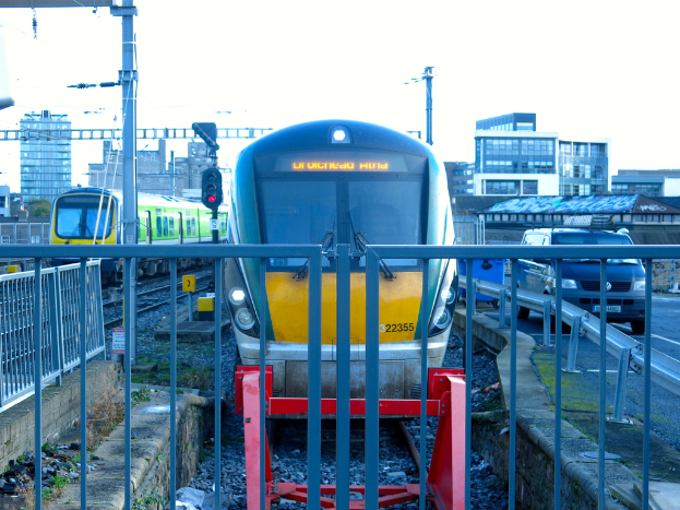 Zwei Züge auf parallelen Gleisen mit einem Zaun vorne, ein Auto auf der Straße rechts, eine Signalleuchte auf einem Pfahl und Gebäude im Hintergrund unter einem Himmel.