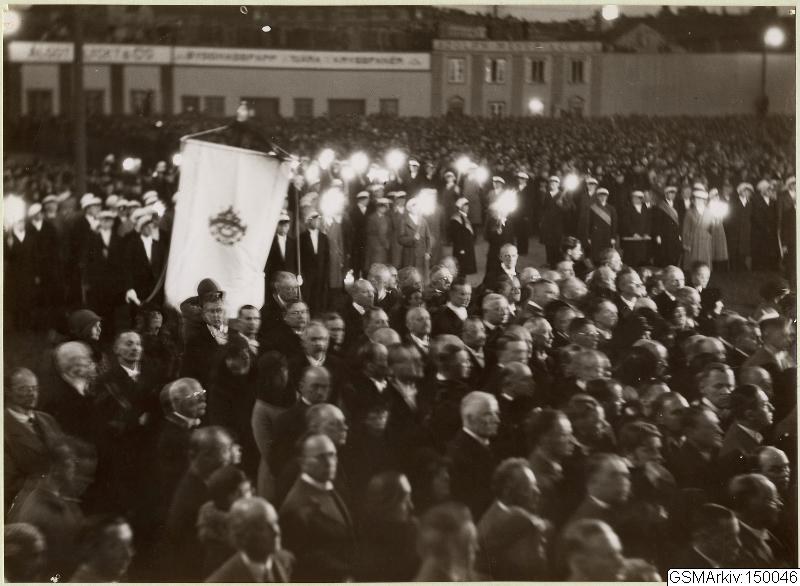 Schwarzes und weißes Foto einer großen Menschenmenge vor einem Gebäude, mit einer Person, die eine Fahne hält, Polen und Lichter im Hintergrund und dem Text "Gustav Klimts Rede an der Berliner Mauer" unten.