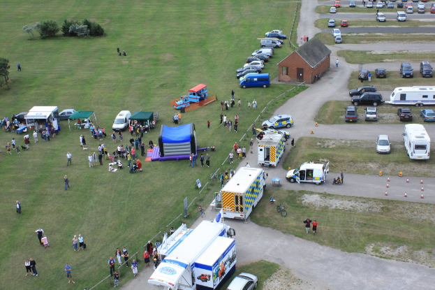 Eine Vogelperspektive einer Szene mit vielen geparkten Fahrzeugen, einigen Menschen in der Nähe von Zelten, einer zentralen Hütte und einigen Bäumen auf der linken Seite.