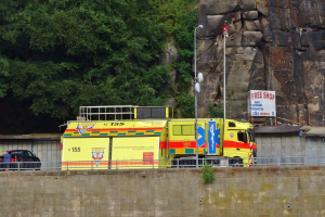 Rettungswagen am Straßenrand neben einer Klippe geparkt, mit einer Wand, einem Geländer, Pfosten, Lampen, Schildern mit Text und anderen Gegenständen im Vordergrund und Bäumen und einem Auto im Hintergrund.
