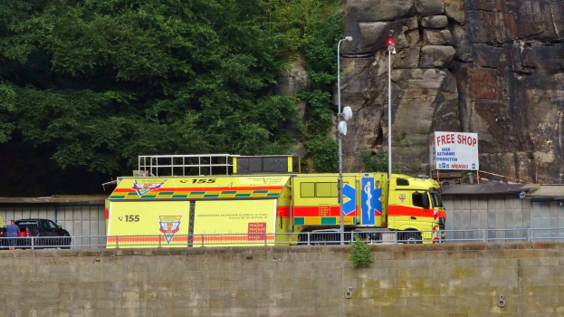Rettungswagen am Straßenrand neben einer Klippe geparkt, mit einer Wand, einem Geländer, Pfosten, Lampen, Schildern mit Text und anderen Gegenständen im Vordergrund und Bäumen und einem Auto im Hintergrund.