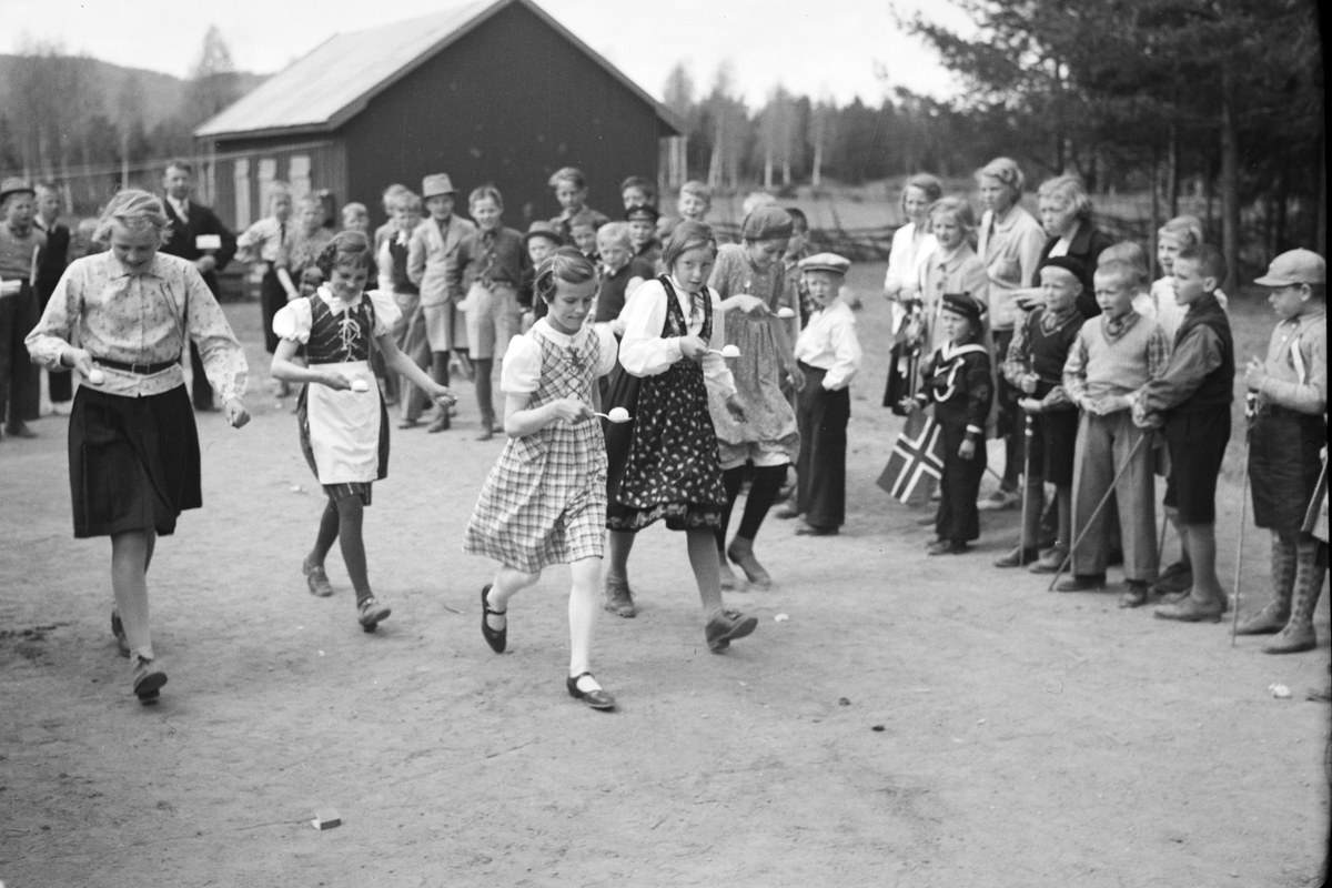 Schwarzes Bild von Kindern, die vor einer Menge mit Fahnen Fußball spielen, mit Bäumen und einem Haus im Hintergrund.