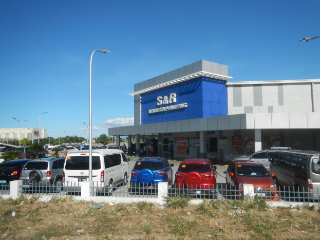 Ein Autohaus mit vorne geparkten Fahrzeugen, umgeben von einem Metallzaun, Gras, Pflanzen, Bäumen, Straßenlaternen und einem bewölkten Himmel.