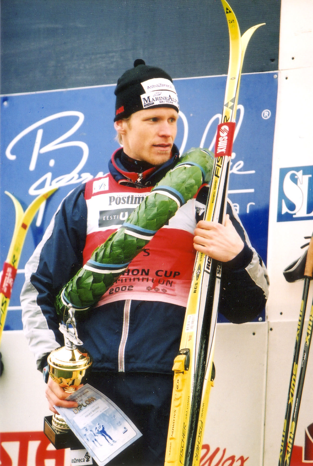 Ein Mann steht auf einem Podium und hält in einer Hand ein Paar Skier und in der anderen eine Trophäe, trägt eine Jacke und eine Mütze und hat ein Schild im Hintergrund.