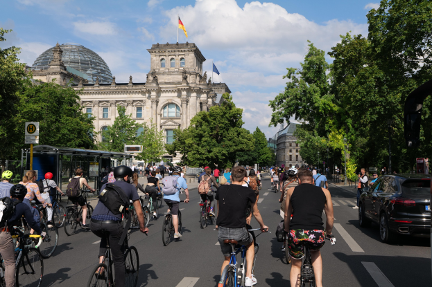 Eine Gruppe von Menschen, die auf Fahrrádern eine von Bäumen gesäumte Straße in Berlin, Deutschland, entlangfahren, mit Gebäuden und einer Bushaltestelle auf der rechten Seite.