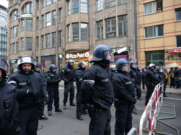 Gruppe von Polizeibeamten hinter Absperrungen vor einer Menge auf einer Straße mit Gebäuden, Schildern, Laternen und einem klaren blauen Himmel im Hintergrund.