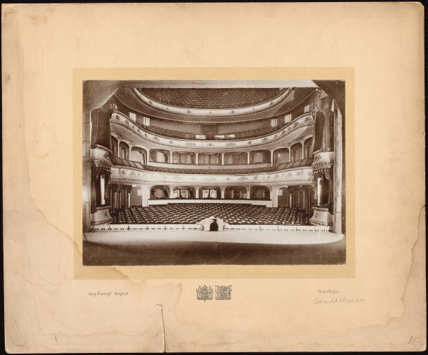 Altes Schwarz-Weiß-Foto des Auditoriums des Royal Albert Hall in London, das die Bühne, Reihen von Stühlen, Säulen, Geländer und Text mit einem Logo unten auf einem dunklen Hintergrund zeigt.