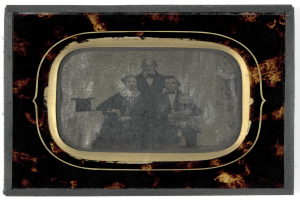 Schwarz-weißes Foto eines Mannes im Anzug und Krawatte und einer Frau in unbekannter Kleidung, die vor einem Fernseher stehen und beide ernst schauen.