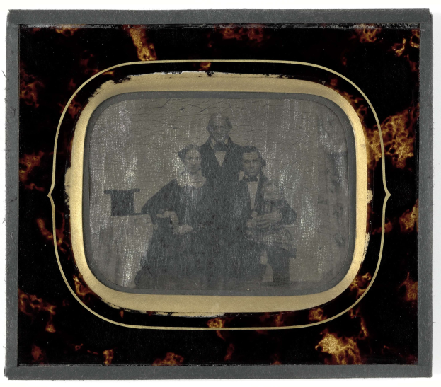 Schwarz-weißes Foto eines Mannes im Anzug und Krawatte und einer Frau in unbekannter Kleidung, die vor einem Fernseher stehen und beide ernst schauen.