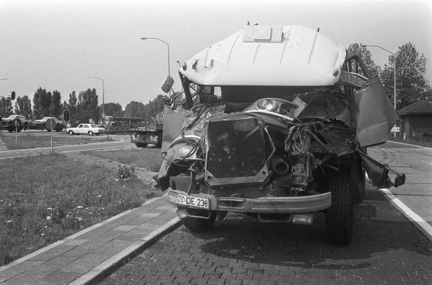 Ein Schwarz-Weiß-Foto eines schwer beschädigten Autos am Straßenrand mit eingedellter Motorhaube und zersplitterten Fenstern sowie anderen Fahrzeugen, Laternenpfählen, Bäumen und einem Haus im Hintergrund.