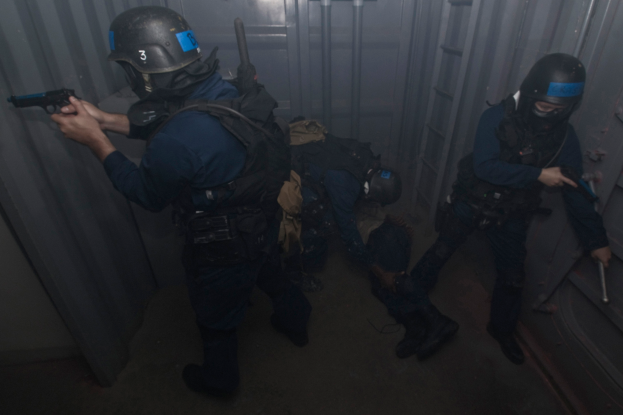 Eine Gruppe von Polizisten in Schutzausrüstung steht zusammen, einer hält eine Waffe, mit einem Metallcontainer und einer Leiter im Hintergrund.