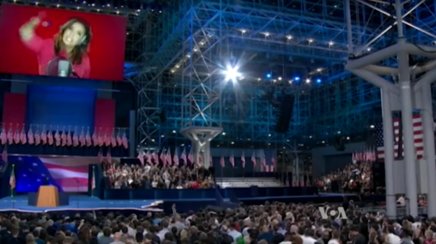 Eine große Menschenmenge sitzt vor einer Bühne mit einem Rednerpult, einem Mikrofon, Flaggen, einem Bildschirm mit dem Bild einer Frau und Licht- und Stativausrüstung, wahrscheinlich bei einer Präsidentschaftsdebatte.