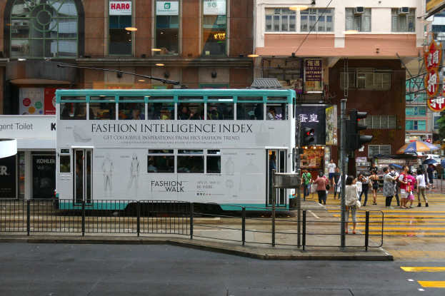 Ein Doppeldeckerbus fährt eine Straße mit hohen Gebäuden entlang, mit Menschen auf dem Gehweg, einige mit Schirmen, Verkehrsampeln, einem Geländer und Schildern mit Text, unter einem klaren blauen Himmel mit Bäumen im Hintergrund.
