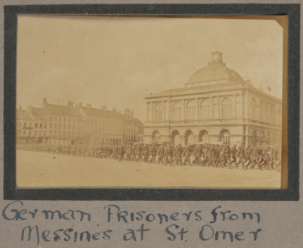 Eine Gruppe deutscher Kriegsgefangener aus Messines steht vor einem Gebäude unter einem klaren blauen Himmel, mit Text am unteren Rand des Bildes.