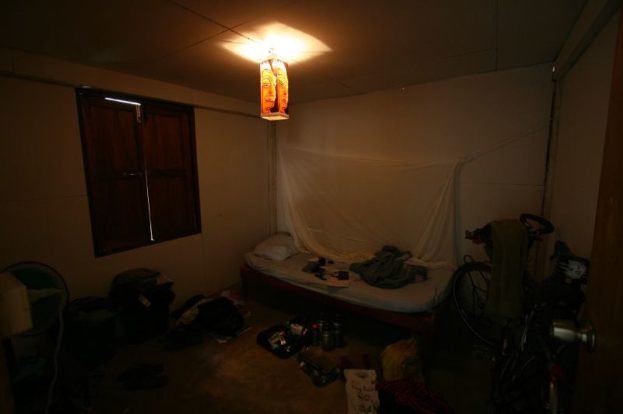 Ein Raum mit einem Bett in der Mitte, ein Fahrrad auf der rechten Seite, ein Fenster auf der linken Seite, ein Tischventilator und Taschen auf der linken Seite und Flaschen unten, mit Licht von oben.