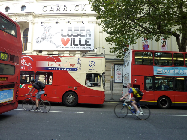 Mehrere Busse und Fahrradfahrer auf einer Straße, mit einem Haus mit Plakaten und einem Baum im Hintergrund.