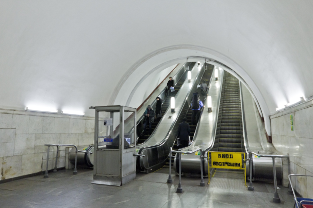 Rolltreppe in einer U-Bahn-Station mit Menschen darauf, umgeben von Geländern, Informationsschildern, Wandlampen und anderen Gegenständen, mit einem Einkaufswagen unten.