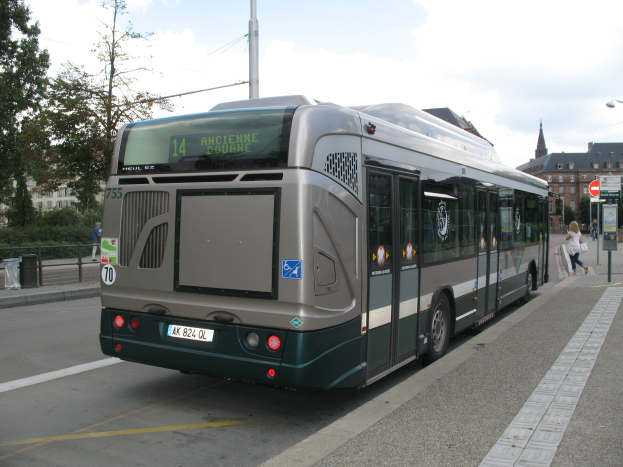 Eine Stadtbus fährt auf einer Straße mit hohen Gebäuden entlang, mit Menschen auf einem Gehweg und Schildern auf Pfählen auf der rechten Seite und Bäumen, Gebäuden und Himmel im Hintergrund.