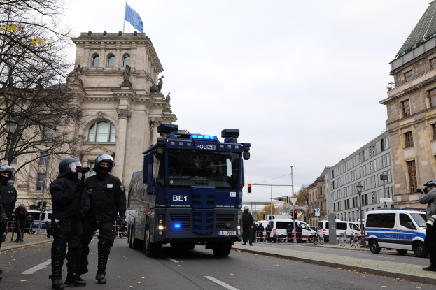 Eine Gruppe von Polizisten in Helmen und mit Waffen in der Hand steht vor einem Gebäude mit Fenstern, Säulen, Bögen und einer Flagge, mit Fahrzeugen auf der Straße, Bäumen, Laternenmasten und einem bewölkten Himmel im Hintergrund.