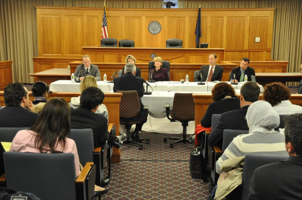 Ein Versammlungsraum mit einer Gruppe von Menschen, zwei ausländischen Flaggen, einem orangen Tisch mit Wasserflaschen und einem Mikrofon.