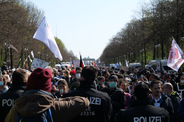 Eine große Gruppe von Menschen steht vor einer Reihe von Polizeibeamten, einige tragen Mützen und Masken, während sie auf einer Demonstration in Berlin, Deutschland, mit Schildern, Fahnen, Laternenpfählen, Bäumen, Fahrzeugen, einem Gebäude und dem Himmel im Hintergrund demonstrieren.