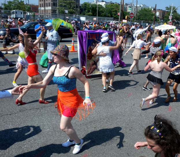 Eine Gruppe von Menschen marschiert durch eine Straße, einige tragen Kappen und halten Schirme, andere tanzen, mit einem Banner, Verkehrskegeln, Fahrzeugen, Laternenmästen, Schildern, Bäumen, Gebäuden und einem klaren blauen Himmel im Hintergrund.