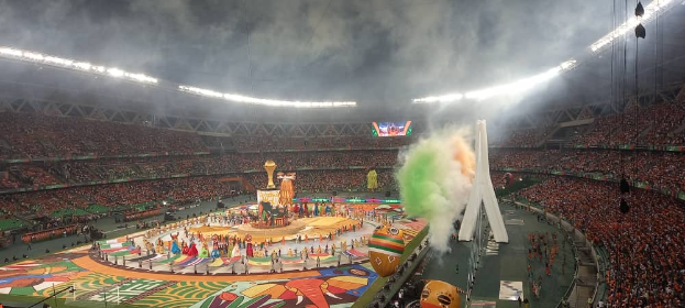 Ein großes Stadion mit Zuschauern, die ein Fußballspiel verfolgen, mit einer zentralen Bühne mit Menschen, Fahnen und Gegenständen, umgeben von Sitzgruppen auf beiden Seiten, mit Lichtern, einem Bildschirm und Rauch an der Spitze.