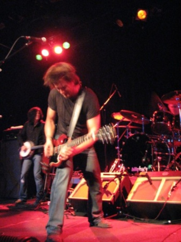 Ein Mann in einem schwarzen Hemd spielt Gitarre, während ein anderer Mann links davon Geige spielt, mit verschiedenen Musikinstrumenten und einer roten Lampe an der Decke im Hintergrund.