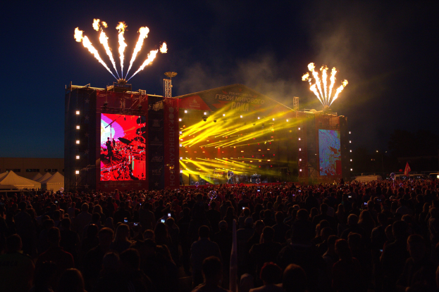 Große Menschenmenge auf einem Musikfestival mit beleuchteter Bühne mit Musikern, umgeben von Bannern, Zelten links und Feuerwerk am dunklen Himmel.