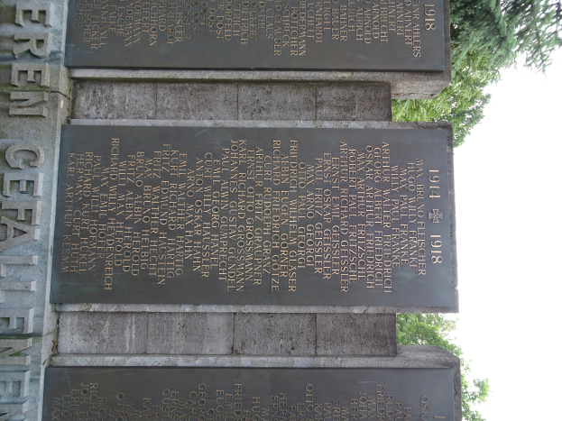 Ein Denkmal mit drei Tafeln vor einem Gebäude, umgeben von Bäumen unter einem klaren blauen Himmel, wahrscheinlich zum Gedenken an Weltkriegsopfer.