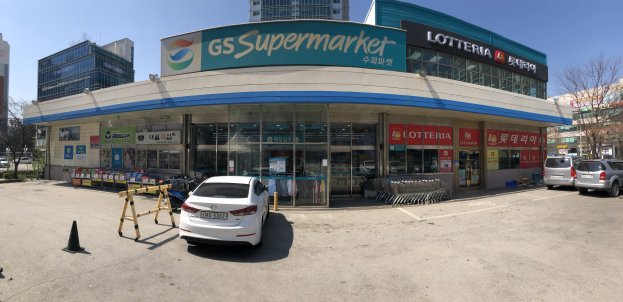 Ein Auto vor einem Laden mit einem "GS Supermarkt"-Schild geparkt, umgeben von anderen Fahrzeugen, Verkehrskegeln und verstreuten Gegenständen, mit Gebäuden, Bäumen und einem klaren blauen Himmel im Hintergrund.