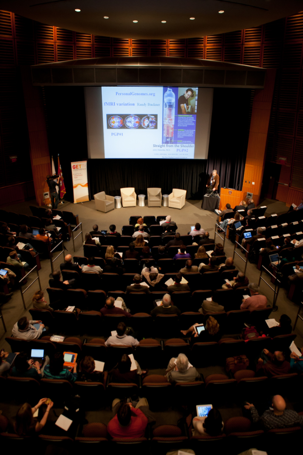 Ein mit Sitzenden Menschen gefüllter Saal, beleuchtet von zahlreichen Lichtern, mit Fahnen links, einem Podium rechts und einem Werbeplakat links.
