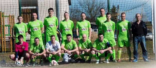 Gruppe von Männern in grünen T-Shirts, die auf einem Fußballfeld stehen oder knien, mit einem Ball und einem Torpfosten, Bäumen und Gebäuden im Hintergrund.
