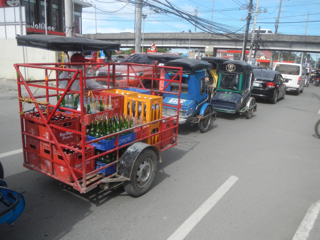 Eine Gruppe von Fahrzeugen auf einer Straße, einschließlich eines Tuk-Tuks mit einem Karren voller Flaschen, einem Gebäude mit Text auf der linken Seite und im Hintergrund Strommasten mit Drähten, eine Brücke, Bäume und einen bewölkten Himmel.