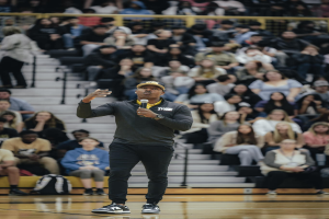 Ein Mann in einer Mütze hält ein Mikrofon auf einem Basketballfeld, mit einer Gruppe von Menschen auf einer Treppe und Taschen auf dem Boden dahinter.