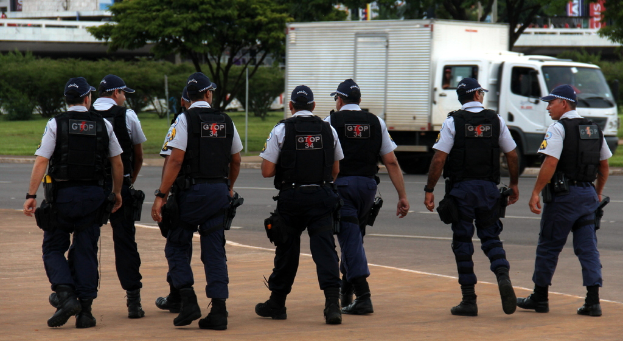 Eine Gruppe von Uniformierten mit Mützen, Hemden, Schutzwesten, Uhren, Hosen und Schuhen, die auf einer Straße mit einem geparkten Lkw auf der rechten Seite neben einem Zaun, Gebäuden, Bäumen, Pflanzen, Polen und Bannern im Hintergrund und einer Brücke im oberen rechten Eck gehen.