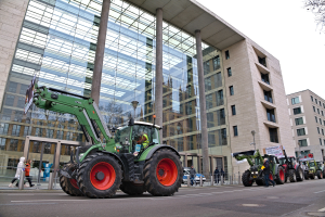 Eine Gruppe von Traktoren fährt auf einer Straße vor einem Gebäude, während Menschen auf dem Gehweg gehen und ein Baum auf der rechten Seite zu sehen ist, während es scheint, dass eine Demonstration stattfindet.