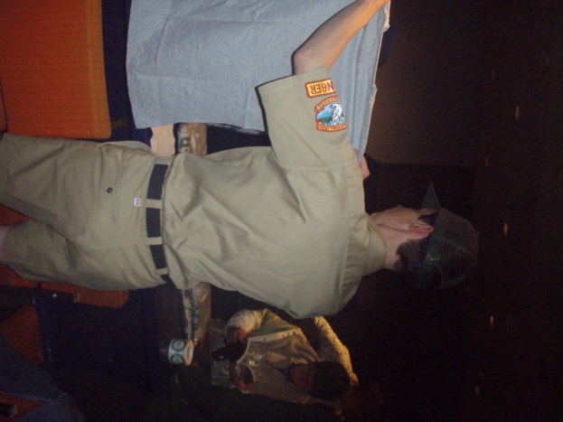 Ein Polizeibeamter hält ein T-Shirt in den Vordergrund, während im Hintergrund ein Junge ein Buch und einen Becher hält und neben einem Schreibtisch und Lampen steht.