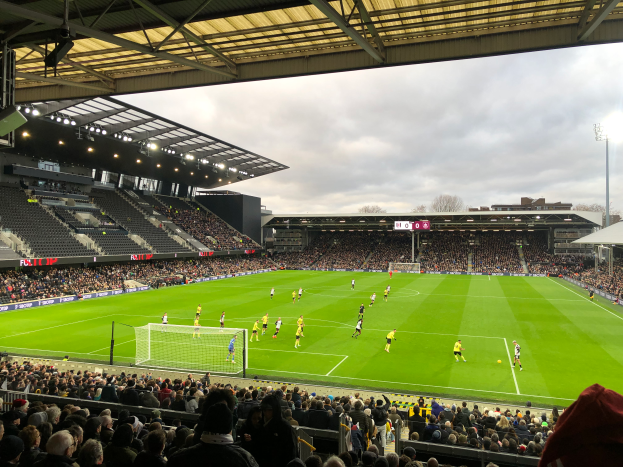 Ein Fußballspiel wird in einem großen Stadion mit Zuschauern in den Rängen, beleuchtet von Deckenlampen, und einem klaren blauen Himmel mit Bäumen und Gebäuden im Hintergrund gespielt.