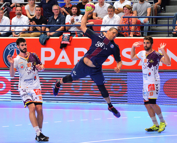 Eine Gruppe von Männern, die Handball auf einem Court spielen, mit einem Ball in der Mitte, Zuschauern im Hintergrund und einem Banner mit der Aufschrift "Futsal-Weltmeisterschaft 2015 - Paris Saint-Germain gegen Olympique Lyon", mit Kleidung, die über das Court verstreut ist.
