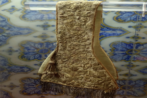 Ein goldener Umhang mit einem detaillierten Muster aus kleinen Kreisen und Linien, der in einer Glasvitrine an einer neutralfarbenen Wand hängt.