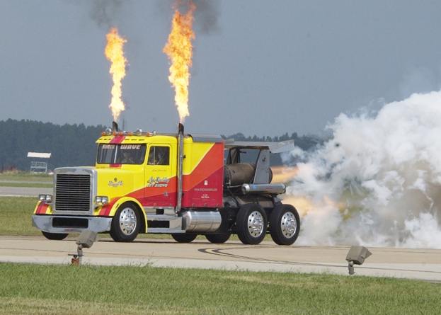 Ein gelber und roter Lkw mit Flammen am Heck fährt auf einer von Gras und Bäumen umgebenen Straße, mit einem klaren blauen Himmel im Hintergrund.