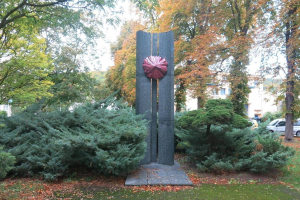 Große Metallskulptur inmitten eines Parks mit Bäumen, Pflanzen und verstreuten Blättern, umgeben von einer Straße mit Fahrzeugen, Gebäuden im Hintergrund und einem klaren blauen Himmel.