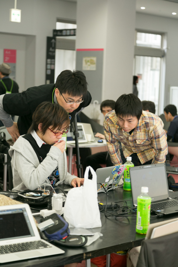 Eine Person sitzt an einem Laptop, während eine andere Person in einem schwarzen T-Shirt hinter ihr steht und ihr über die Schulter schaut.