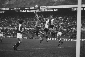 Eine Gruppe von Männern, die auf einem Feld Fußball spielen, mit einem Torpfosten auf der rechten Seite und einer Zuschauermenge im Hintergrund, dargestellt in Schwarz-Weiß.