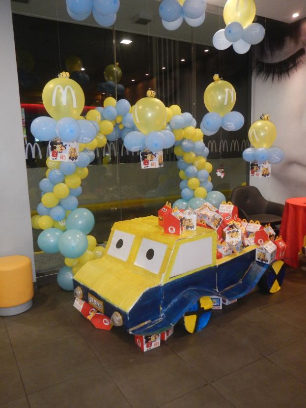 Eine McDonald's-Geburtstagsparty-Szene mit einem gelben und blauen Lastwagen, der mit Ballons geschmückt ist, einem rot gedeckten Tisch mit Gegenständen, Hockern, einer Glaswand und Deckenleuchten.
