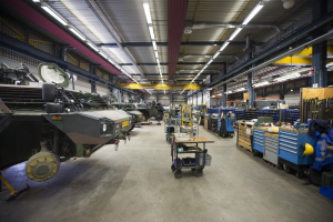 Großer Lagerinnenraum mit Maschinen, Fahrzeugen auf der linken Seite, Transportwagen und Holzboxen auf der rechten Seite und Deckenbeleuchtung.